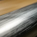 Un primo piano macro di un laminato ibrido alluminio-legno, 100mm Macro lens, illuminazione da studio controllata, che mostra la texture del legno di betulla e la lucentezza dell'alluminio, con una leggera curvatura che suggerisce flessibilità e resistenza, per strutture leggere sostenibili.