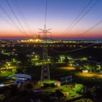 Veduta aerea di un paesaggio energetico eterogeneo in Mozambico al tramonto, che mostra una centrale a gas illuminata in lontananza, linee elettriche che attraversano il territorio e piccole comunità rurali con alcune luci sparse da sistemi solari off-grid. Obiettivo grandangolare 10mm, lunga esposizione per catturare le luci e il cielo, forte contrasto tra sviluppo industriale e aree rurali.