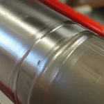 Primo piano di una giunzione tra due tubi di acciaio per oleodotto realizzata con saldatura meccanica allo stato solido. Dettaglio macro con focale 100mm, illuminazione controllata che evidenzia la finitura metallica e la zona di contatto ad alta pressione. Alta definizione, messa a fuoco precisa.