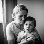 Ritratto fotografico toccante di una madre visibilmente stressata che cerca di consolare un bambino piccolo (3-4 anni) durante il lockdown pandemico. Ambiente domestico, luce naturale dalla finestra, obiettivo prime 50mm, profondità di campo che isola la coppia, toni leggermente desaturati, bianco e nero film.