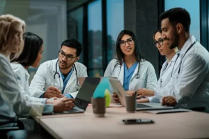 Un gruppo di giovani medici specializzandi, uomini e donne di diverse etnie, discute animatamente attorno a un tavolo luminoso in una moderna sala riunioni di un ospedale, alcuni consultano tablet e laptop. Fotografia di ritratto, obiettivo da 35mm, profondità di campo, luce naturale soffusa che entra da grandi finestre, atmosfera collaborativa e dinamica.