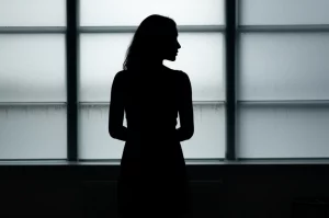 Immagine concettuale di una silhouette di donna pensierosa che guarda fuori da una finestra in un ambiente ospedaliero, con un leggero effetto controluce. Obiettivo prime 50mm, profondità di campo, toni freddi per trasmettere introspezione e la serietà dell'argomento trattato, come l'aborto interno e la gestione farmacologica.