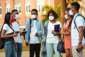 Immagine fotorealistica di studenti universitari ghanesi in un campus, che discutono animatamente ma a distanza, alcuni con mascherine. La scena cattura la dinamica sociale giovanile durante la pandemia. Obiettivo da 35mm, luce del tardo pomeriggio, colori caldi e leggero effetto filmico per un'atmosfera coinvolgente e autentica.