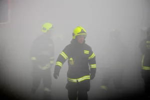 Immagine fotorealistica di un gruppo di vigili del fuoco in equipaggiamento completo che emergono da una densa nebbia di fumo, con un focus selettivo su uno di loro in primo piano, volto segnato dalla fatica ma determinato. Telephoto zoom 100-400mm, fast shutter speed, action tracking, luce drammatica che filtra attraverso il fumo.