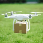 Primo piano di un drone per consegne moderno e bianco con un pacco agganciato, in volo stazionario sopra un prato verde lussureggiante, sfondo leggermente sfocato (bokeh), obiettivo prime 50mm, luce diurna naturale, alta definizione.
