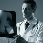 Ritratto di un medico oncologo con camice bianco, pensieroso, mentre osserva una radiografia di un rene con aree metastatiche evidenziate. L'illuminazione è drammatica, in stile film noir, con un duotone blu e grigio per accentuare la serietà della situazione. Obiettivo da 35mm, profondità di campo ridotta per mettere a fuoco il volto del medico e la lastra, simboleggiando la difficile scelta terapeutica nel carcinoma renale metastatico.