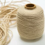 Macro fotografia, 105mm lens, di un fascio di fibre di canapa finemente lavorate, simili al cotone, accanto a un rocchetto di filato misto canapa-cotone. Alta definizione, messa a fuoco precisa, illuminazione controllata da studio, che enfatizza la trasformazione della materia prima in prodotto tessile.