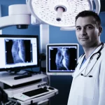 Primo piano di un radiologo interventista che esegue un'embolizzazione dell'arteria bronchiale in una sala angiografica high-tech. L'immagine, con focale 35mm e un leggero effetto duotone blu e grigio, cattura la concentrazione del medico e la precisione della procedura, con monitor che mostrano immagini angiografiche dei vasi polmonari.