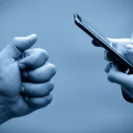 Immagine simbolica: una mano di un genitore che cerca di raggiungere la mano di un adolescente immerso nella luce bluastra di uno schermo di smartphone, obiettivo prime 50mm, profondità di campo, toni blu e grigi duotone, focus sulla distanza tra le mani.