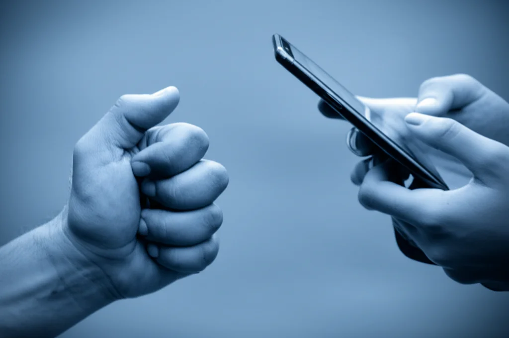 Immagine simbolica: una mano di un genitore che cerca di raggiungere la mano di un adolescente immerso nella luce bluastra di uno schermo di smartphone, obiettivo prime 50mm, profondità di campo, toni blu e grigi duotone, focus sulla distanza tra le mani.