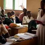 Un'aula scolastica nigeriana, studenti attenti, un insegnante sembra perplesso di fronte a dei documenti di valutazione. Luce naturale dalla finestra, atmosfera di studio ma con un velo di incertezza. Prime lens, 35mm, depth of field, colori caldi e leggermente desaturati.