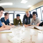 wide-angle, un'aula scolastica moderna e luminosa, con un gruppo eterogeneo di studenti adolescenti (maschi e femmine, diverse etnie) seduti attorno a un grande tavolo, attivamente impegnati nell'esaminare e discutere quaderni di progettazione ingegneristica e alcuni piccoli prototipi sparsi sul tavolo, l'atmosfera è collaborativa e concentrata, obiettivo grandangolare da 24mm, messa a fuoco nitida su tutto il gruppo, luce naturale abbondante che entra da ampie finestre.