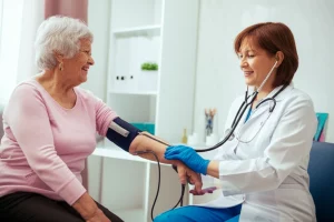 Primo piano di un medico che misura la pressione sanguigna a una donna anziana sorridente in un ambulatorio luminoso. Obiettivo prime 35mm, profondità di campo per enfatizzare i soggetti, toni caldi e accoglienti.