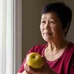 Ritratto fotografico realistico di una donna cinese di mezza età (circa 55 anni) con diabete di tipo 2, seduta vicino a una finestra con luce naturale soffusa. Ha un'espressione pensierosa ma serena, forse mentre controlla il suo misuratore di glicemia o tiene in mano una mela. Lente prime da 50mm, profondità di campo ridotta per sfocare leggermente lo sfondo interno (una casa accogliente ma semplice), mettendo in risalto la persona. Illuminazione controllata per un effetto morbido e intimo.