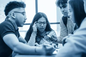 Un gruppo eterogeneo di giovani adolescenti, alcuni con tratti somatici Latinx, discutono animatamente in un ambiente scolastico o ricreativo. Una sigaretta elettronica è visibile ma non al centro dell'attenzione, magari appoggiata su un tavolo o tenuta distrattamente. Obiettivo da ritratto 50mm, luce naturale che entra da una finestra, effetto bokeh per sfocare leggermente lo sfondo ed enfatizzare i volti e le espressioni dei ragazzi, duotone blu e grigio per un'atmosfera seria ma coinvolgente.