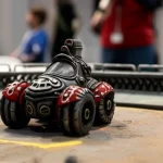 Primo piano di una miniatura di un veicolo fantascientifico, finemente dettagliata, al centro di un'arena di gara in miniatura. L'immagine è scattata con un obiettivo macro da 100mm per esaltare i dettagli, con illuminazione da studio che crea ombre drammatiche e riflessi metallici. Sullo sfondo, sfocati, si intravedono altri partecipanti e giudici, suggerendo l'atmosfera di un contest STEAM. Alta definizione e messa a fuoco precisa sul modello.