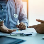Un medico specialista in ematologia discute con un paziente adulto i risultati di un trattamento innovativo per la trombocitopenia immune (ITP), in un ambiente clinico moderno e luminoso. Primo piano sulle mani del medico che indicano un grafico, obiettivo 50mm, profondità di campo.