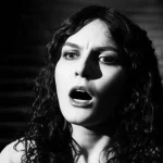 Una potente immagine teatrale di Medea, divisa tra rabbia e disperazione, che simboleggia la lotta contro la xenofobia e la misoginia. Primo piano del volto di un'attrice che interpreta Medea, illuminazione drammatica con forti contrasti, obiettivo prime 35mm, effetto film noir per accentuare l'intensità emotiva. Sullo sfondo, elementi scenici che richiamano sia la sua origine straniera sia il rifiuto subito.