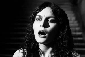 Una potente immagine teatrale di Medea, divisa tra rabbia e disperazione, che simboleggia la lotta contro la xenofobia e la misoginia. Primo piano del volto di un'attrice che interpreta Medea, illuminazione drammatica con forti contrasti, obiettivo prime 35mm, effetto film noir per accentuare l'intensità emotiva. Sullo sfondo, elementi scenici che richiamano sia la sua origine straniera sia il rifiuto subito.