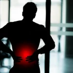 Immagine concettuale che mostra la silhouette di un lavoratore (impiegato d'ufficio) con la zona lombare evidenziata in rosso acceso a simboleggiare il dolore, su uno sfondo sfocato di un moderno open space. Fotografia simbolica, obiettivo 50mm, profondità di campo, colori neutri con forte contrasto sul rosso.