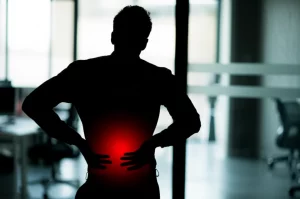 Immagine concettuale che mostra la silhouette di un lavoratore (impiegato d'ufficio) con la zona lombare evidenziata in rosso acceso a simboleggiare il dolore, su uno sfondo sfocato di un moderno open space. Fotografia simbolica, obiettivo 50mm, profondità di campo, colori neutri con forte contrasto sul rosso.