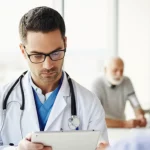 Immagine fotorealistica di un medico che esamina i risultati di un esame del sangue su un tablet in un ambiente ospedaliero moderno, con un paziente anziano visibile ma leggermente sfocato sullo sfondo. Obiettivo 35mm, profondità di campo, luce naturale soffusa dalla finestra, espressione concentrata del medico.