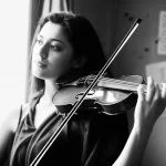 Ritratto fotografico di un musicista classico (violinista donna, 30 anni) che guarda fuori da una finestra con espressione pensierosa, il suo violino appoggiato sulla spalla ma non in posizione di suono, luce naturale laterale che illumina metà del viso, sfondo leggermente sfocato di una stanza con spartiti, profondità di campo ridotta, lente prime 50mm, bianco e nero con leggero contrasto.