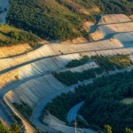Un paesaggio che mostra la difficile coesistenza tra un'area mineraria attiva a cielo aperto e la lussureggiante natura circostante, con un corridoio ecologico visibile che cerca di farsi strada tra le due. Immagine fotorealistica, obiettivo grandangolare 24mm per catturare l'ampiezza della scena, luce del tardo pomeriggio per evidenziare i contrasti e le texture, dettagli nitidi sia sull'attività industriale che sulla vegetazione.