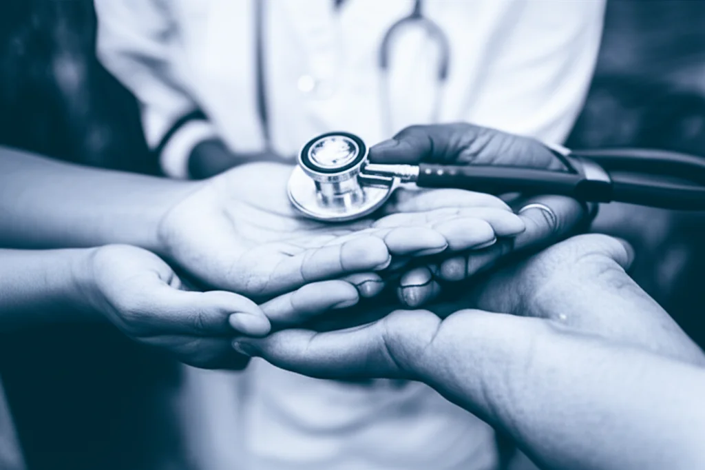 Un primo piano di mani di diversa etnia che si sostengono a vicenda, con sullo sfondo sfocato simboli di cura e salute come un cuore e uno stetoscopio. Prime lens, 35mm, depth of field, duotone blu e grigio per un senso di serietà e speranza.