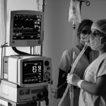 Un paziente in un letto di terapia intensiva indossa una maschera per la ventilazione non invasiva (NIV). Accanto, un moderno ventilatore e monitor con parametri vitali. Un operatore sanitario controlla la situazione. L'immagine è un ritratto, obiettivo 35mm, con profondità di campo per focalizzare sul paziente e la maschera, in bianco e nero per un effetto drammatico ma speranzoso.
