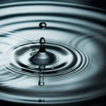 Fotografia macro di una goccia d'acqua cristallina che cade in un bicchiere d'acqua pulita, creando increspature concentriche sulla superficie, illuminazione laterale controllata per esaltare la trasparenza e i riflessi, obiettivo macro 100mm, alta definizione, messa a fuoco precisa sul punto di impatto della goccia.