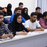 Fotografia di un gruppo eterogeneo di studenti di infermieristica iraniani, maschi e femmine, in un'aula moderna e luminosa, attenti durante una lezione sull'etica professionale. Alcuni prendono appunti, altri ascoltano con interesse. Prime lens 35mm, profondità di campo per mettere a fuoco gli studenti in primo piano, luce naturale che entra dalle finestre.
