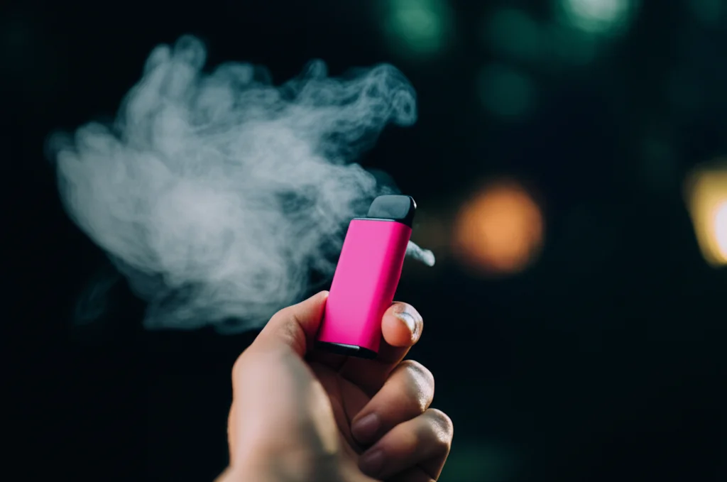 Una mano che tiene una sigaretta elettronica usa e getta colorata, con una leggera nuvola di vapore che si dissolve. Macro lens, 60mm, high detail, precise focusing, con un leggero effetto bokeh sullo sfondo per concentrare l'attenzione sul prodotto e sul vapore.
