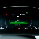 Primo piano del cruscotto digitale di un'auto elettrica moderna, che mostra in modo prominente l'indicatore dello stato di carica (SOC) della batteria come una barra percentuale luminosa e un'autonomia residua in chilometri. Illuminazione ambientale interna soffusa, obiettivo 50mm, messa a fuoco nitida sull'indicatore SOC, sfondo leggermente sfocato (effetto bokeh).