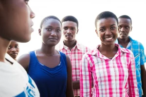 Un gruppo di giovani adulti mozambicani, alcuni con espressioni pensierose altri più speranzose, interagiscono in un contesto comunitario che suggerisce supporto e dialogo. Fotografia di gruppo, obiettivo da 35mm, luce naturale, colori vivaci ma realistici, profondità di campo media per includere il contesto.
