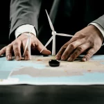 Fotografia concettuale: una mappa stilizzata dell'Asia Centrale su un tavolo di legno scuro. Due mani, una con una manica di giacca europea e l'altra con una manica di stile asiatico/cinese, si contendono simboli di energia (una turbina eolica e una goccia di petrolio) posizionati sulla mappa. Illuminazione drammatica, obiettivo macro da 60mm per dettaglio sui simboli e le mani, leggera profondità di campo.