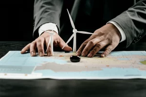 Fotografia concettuale: una mappa stilizzata dell'Asia Centrale su un tavolo di legno scuro. Due mani, una con una manica di giacca europea e l'altra con una manica di stile asiatico/cinese, si contendono simboli di energia (una turbina eolica e una goccia di petrolio) posizionati sulla mappa. Illuminazione drammatica, obiettivo macro da 60mm per dettaglio sui simboli e le mani, leggera profondità di campo.