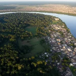 Immagine aerea composita che mostra la fitta foresta amazzonica che sfuma gradualmente in un'area rurale con piccole coltivazioni e poi in una vivace ma caotica area urbana lungo un fiume. Obiettivo grandangolare 18mm, luce del tramonto che crea lunghe ombre, mettendo in evidenza l'interconnessione e la transizione tra i diversi ambienti amazzonici.
