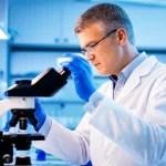 Un ricercatore esperto, in camice bianco, osserva attentamente un campione al microscopio in un laboratorio di ricerca medica avanzato. L'ambiente è illuminato da luci brillanti e pulite, con attrezzature scientifiche moderne visibili sullo sfondo. Prime lens, 50mm, depth of field, l'espressione del ricercatore è concentrata e suggerisce dedizione e speranza nella lotta contro malattie come il mieloma multiplo.