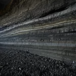 Immagine fotorealistica di una sezione trasversale di una miniera di carbone moderna e profonda, illuminata da fari. Si vedono strati di roccia e carbone sotto stress. Utilizzare un obiettivo grandangolare (10-24mm) per catturare l'ampiezza, con messa a fuoco nitida e illuminazione controllata per evidenziare i dettagli geologici e la tensione implicita. Evocare un senso di profondità e potenziale pericolo.