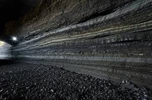 Immagine fotorealistica di una sezione trasversale di una miniera di carbone moderna e profonda, illuminata da fari. Si vedono strati di roccia e carbone sotto stress. Utilizzare un obiettivo grandangolare (10-24mm) per catturare l'ampiezza, con messa a fuoco nitida e illuminazione controllata per evidenziare i dettagli geologici e la tensione implicita. Evocare un senso di profondità e potenziale pericolo.