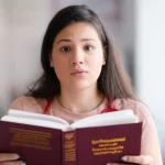 A 35mm portrait of a young medical student looking overwhelmed while holding a thick medical textbook written in a foreign language, depth of field, soft lighting.
