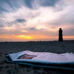 Paesaggio grandangolare, obiettivo da 15 mm. Una giovane persona araba solita si è sgauuetta contro un cielo tramonto drammatico e infuocato, guardando verso l'orizzonte con una posizione contemplativa. In primo piano, leggermente sfigurato su un terreno polveroso, si trova un giornale scartato con titoli di guerra sfocati ma riconoscibili. Un forte senso di contemplazione e resilienza tra le turbolenze globali. Focus acuto sulla silhouette e sul cielo vibrante, lunga esposizione che crea nuvole lisce.