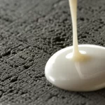 Un campione di calcestruzzo modificato con lattice di gomma naturale, con una goccia di lattice bianco che cade su di esso, illuminazione da studio precisa e controllata, obiettivo macro 90mm, alta definizione della texture superficiale del calcestruzzo e della viscosità del lattice, sfondo neutro.