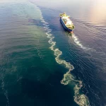 Immagine fotorealistica di un fronte oceanico submesoscala visto dall'alto, con una nave da ricerca scientifica che lo attraversa. L'acqua più calda (leggermente più chiara e con tonalità tendenti al verde acqua) incontra l'acqua più fredda (più scura, blu intenso) creando una linea di demarcazione visibile con piccoli vortici e turbolenze superficiali che increspano l'acqua. Obiettivo zoom teleobiettivo, 200mm, per comprimere la prospettiva e mettere a fuoco i dettagli del fronte. Luce del tardo pomeriggio che crea lunghe ombre e riflessi dorati sull'acqua, cielo parzialmente nuvoloso. Profondità di campo ridotta per enfatizzare il fronte.