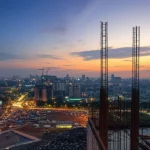 Skyline di Dhaka al tramonto con cantieri edili in primo piano, simboleggiando la rapida urbanizzazione e la sfida dei rifiuti da costruzione. Fotografia di paesaggio urbano, obiettivo grandangolare 18mm, lunga esposizione per nuvole setose e luci della città vibranti, messa a fuoco nitida sull'orizzonte.