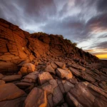 Un'immagine grandangolare di un paesaggio vulcanico australiano antico, con formazioni rocciose suggestive e un cielo drammatico al tramonto, che evoca le antiche storie aborigene. Wide-angle, 10mm, long exposure, sharp focus, colori caldi del tramonto che contrastano con le rocce scure.