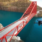 Foto aerea grandangolare (18mm) di un drone Elios 2 che ispeziona da vicino la complessa struttura inferiore di un grande ponte in acciaio. Luce del tardo pomeriggio, messa a fuoco nitida sulla struttura del ponte e sul drone, lunga esposizione per sfocare leggermente l'acqua sottostante, simboleggiando l'ispezione avanzata delle infrastrutture.