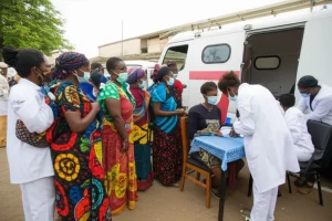 Fotografia grandangolare di una clinica mobile per vaccinazioni allestita in una piazza di una città tanzaniana, con una piccola folla di persone in attesa ordinata e personale sanitario in camice bianco che somministra vaccini. Obiettivo grandangolare 24mm, luce diurna brillante, colori vivaci degli abiti e dell'ambiente, messa a fuoco nitida su tutta la scena per mostrare l'attività comunitaria e l'intervento di salute pubblica.