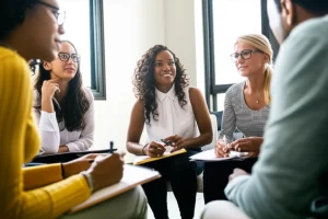 Un gruppo multietnico di persone (Nere e Latine in primo piano) in cerchio, che collaborano e discutono animatamente durante un incontro di un community advisory board. Stile fotorealistico, obiettivo da 35mm, luce naturale che entra da una finestra laterale, espressioni concentrate e partecipative, alcuni con quaderni e penne. Ambientazione: una sala comunitaria semplice ma accogliente.
